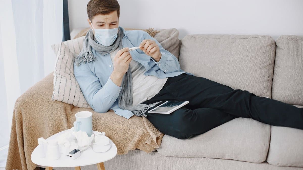 Man wearing a mask checks a thermometer while resting on a couch, surrounded by tissues.
