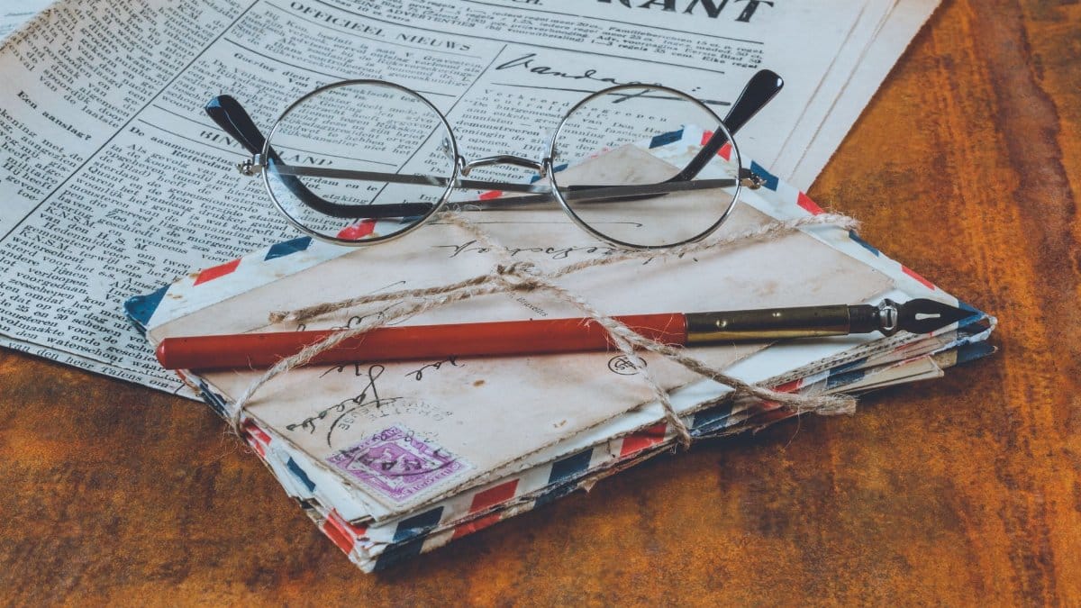 Historical letters and a fountain pen on a vintage wooden table, alongside eyeglasses and a newspaper.