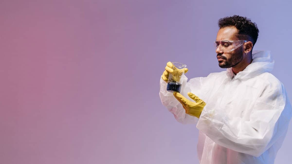 A scientist in protective gear observes a liquid in a beaker under colored lighting.