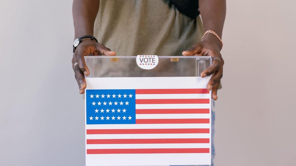 Close-up of a person placing a vote in a transparent ballot box with an American flag print.
