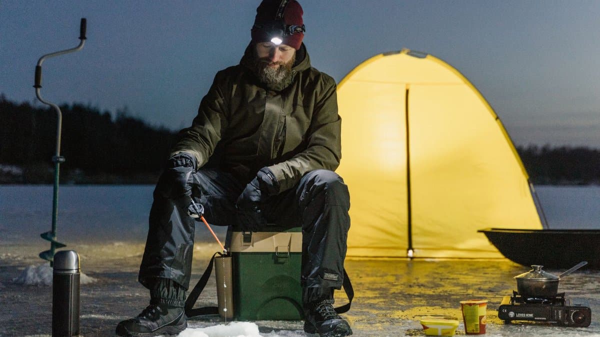 A man ice fishing by a camping tent on a frozen lake at night, embodying winter adventure.