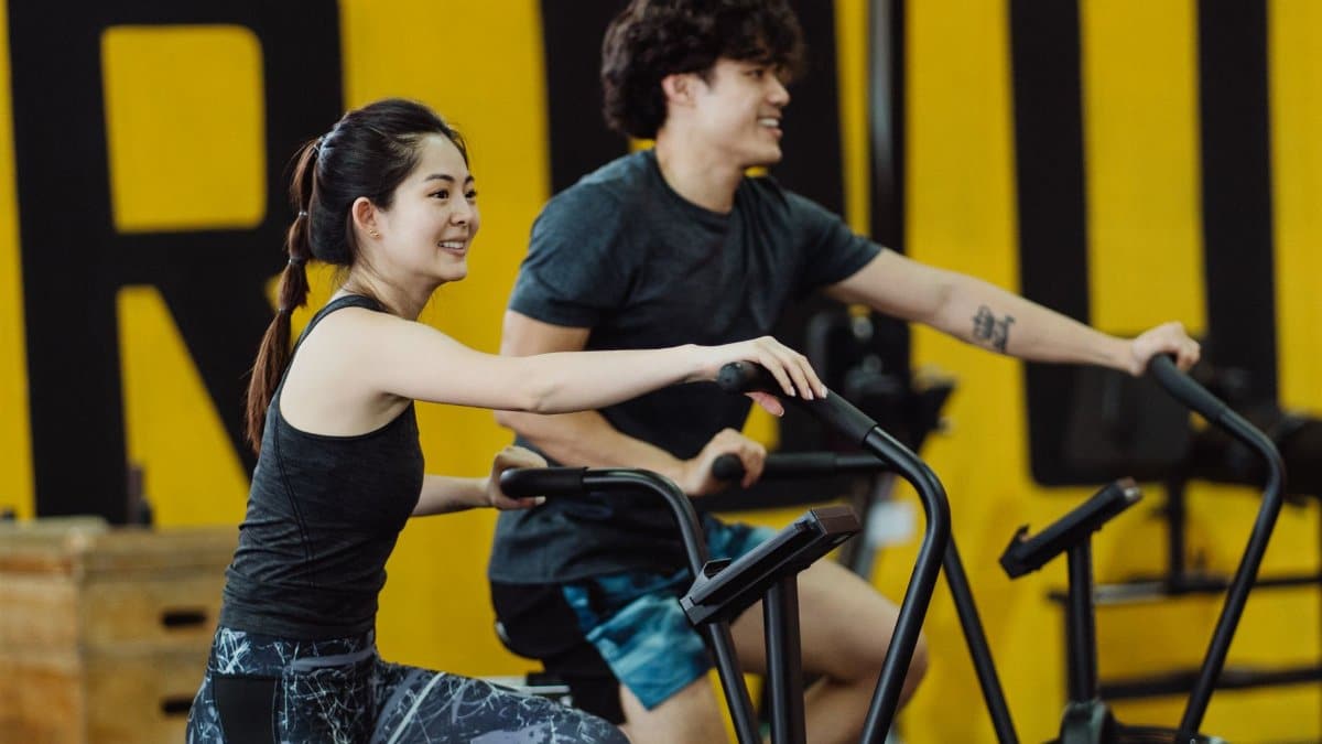 Asian man and woman enjoying a workout on stationary bikes in a gym, smiling and energetic.