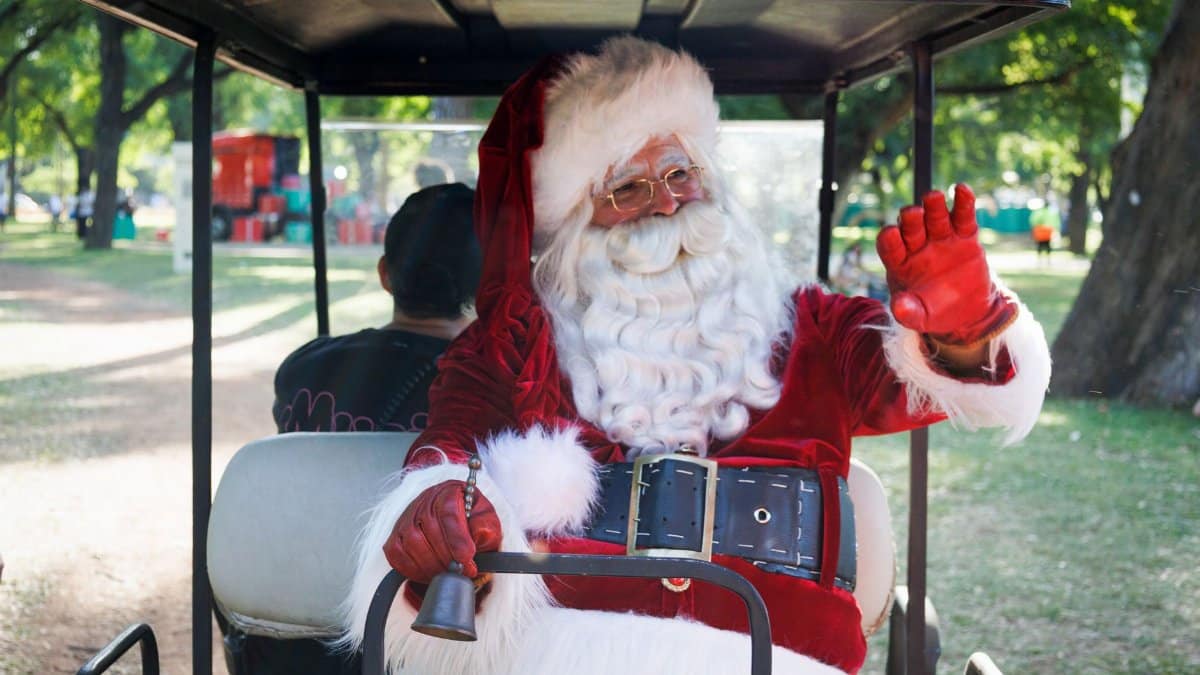 Santa Claus in a golf cart, waving with a bell in hand in a sunny park setting.