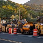 Road construction site with machinery and traffic cones under a scenic hill at sunset.