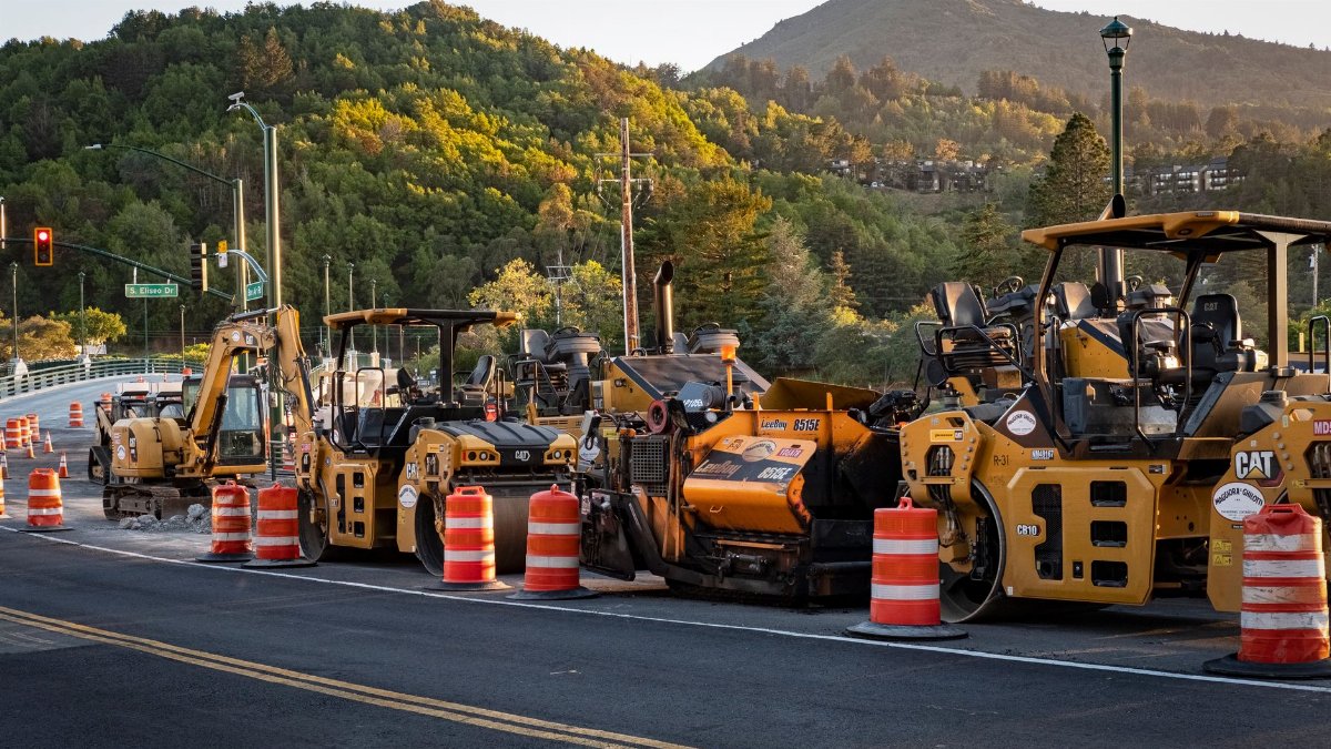 Road construction site with machinery and traffic cones under a scenic hill at sunset.