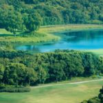 Aerial view of a lush green golf course surrounded by forest and a tranquil lake.