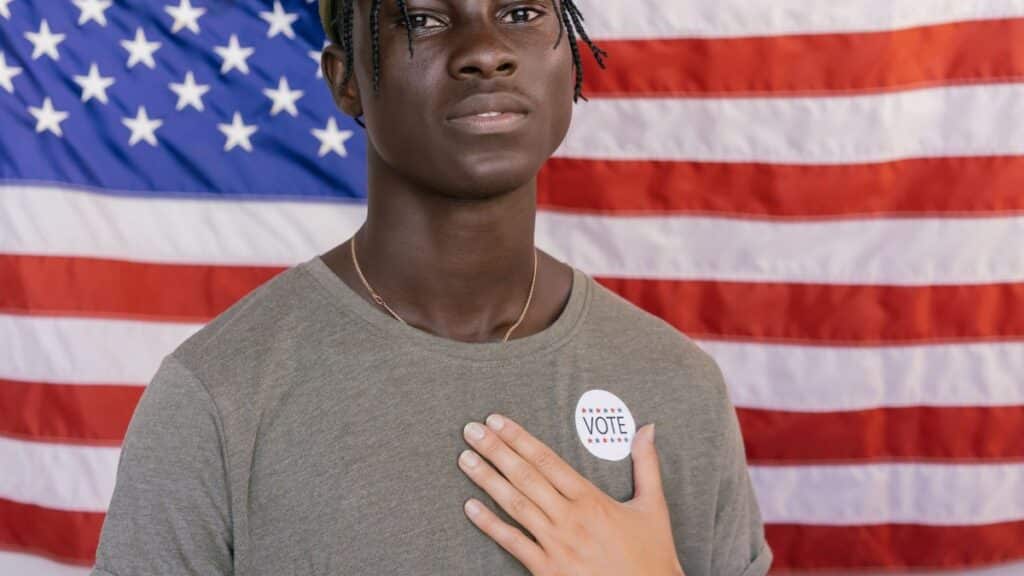 A young adult stands before an American flag wearing a vote badge, symbolizing civic engagement.