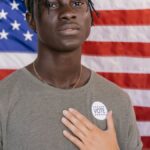 A young adult stands before an American flag wearing a vote badge, symbolizing civic engagement.