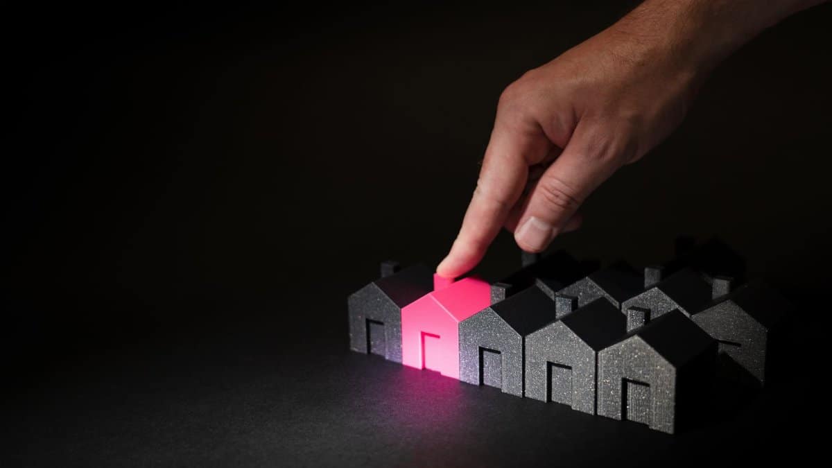 A hand selecting a glowing pink house among dark miniature houses, symbolizing real estate choices.