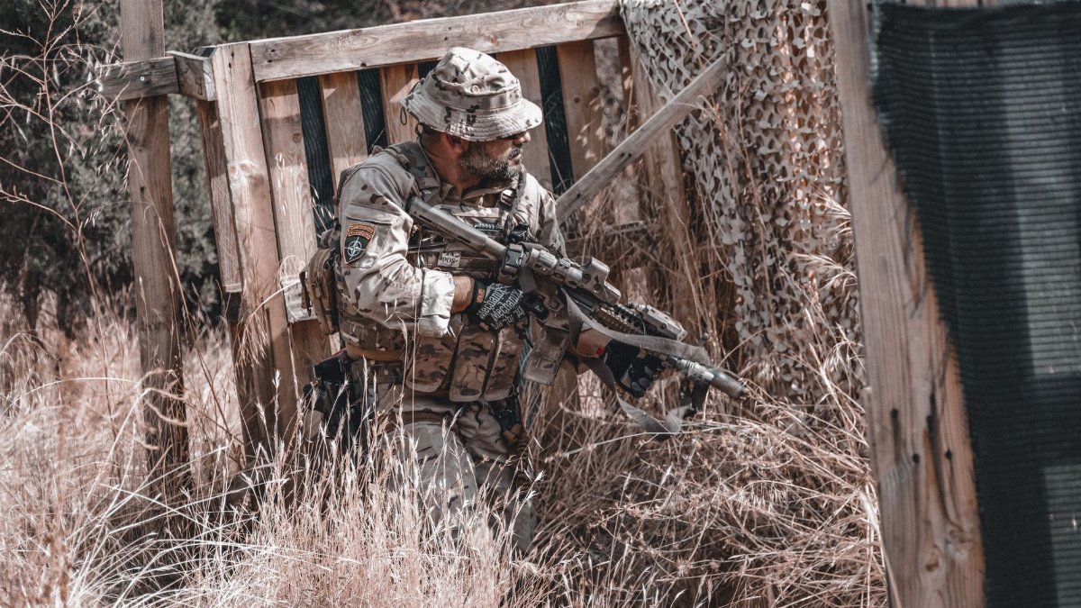 A soldier in full camouflage gear holding a rifle, hiding in an outdoor setting, Spain.
