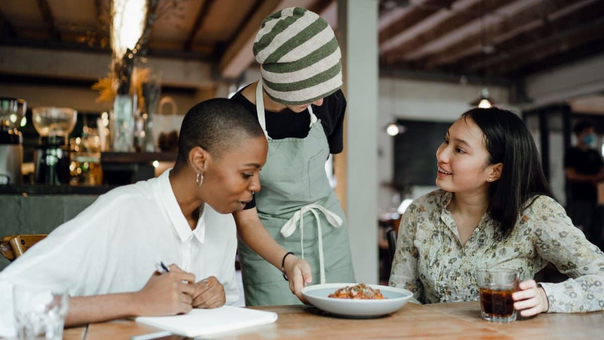 Positive smiling colleagues tasting food from new menu and taking notes while sitting at table in cafe and talking to waitress in uniform