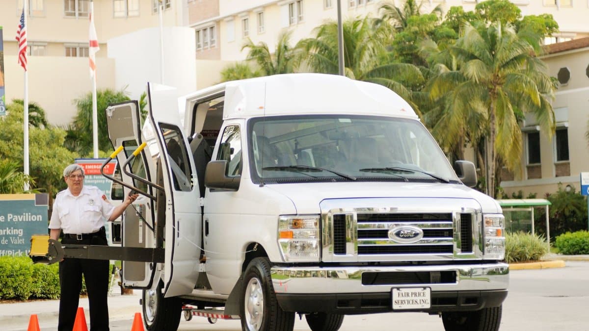 A parking assistant operating a van with a wheelchair lift in a palm-lined parking area.