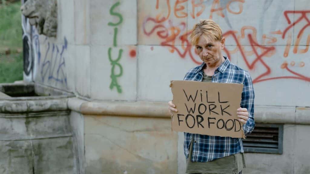 A homeless woman standing outdoors holding a sign that reads 'Will Work For Food'.