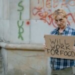 A homeless woman standing outdoors holding a sign that reads 'Will Work For Food'.