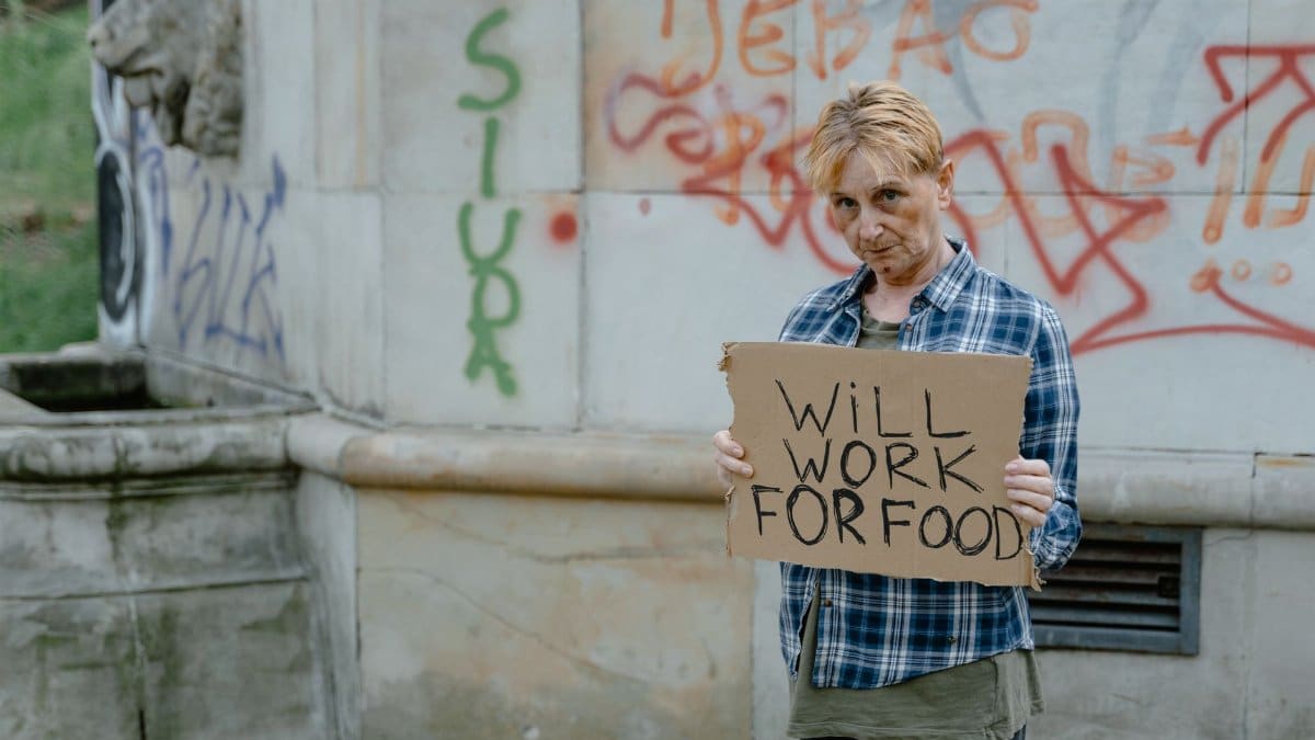 A homeless woman standing outdoors holding a sign that reads 'Will Work For Food'.