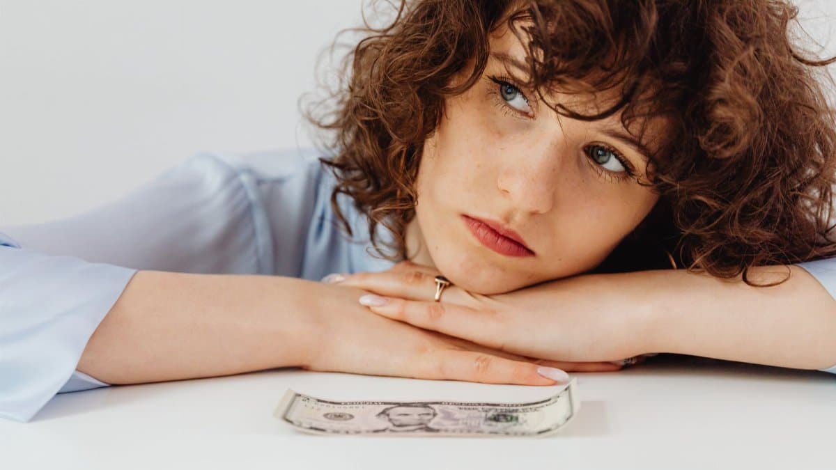 Worried woman with curly hair leaning on white surface with a US dollar bill.