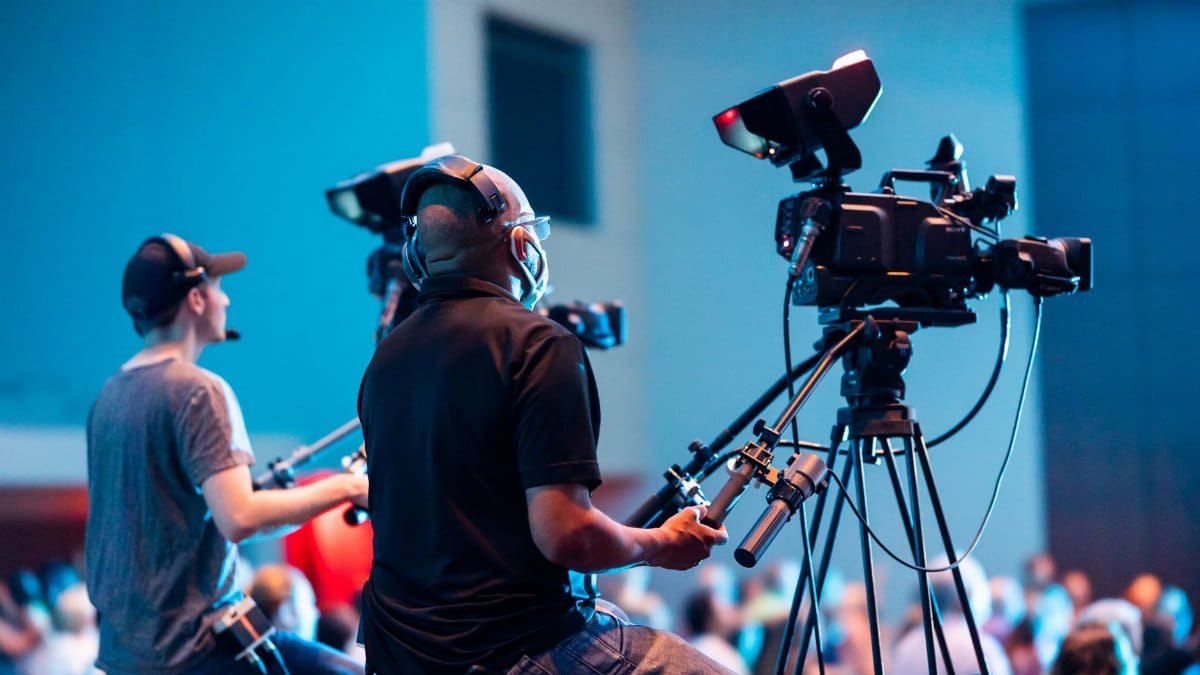 Two cameramen operate professional video cameras at an indoor event with a lively audience.