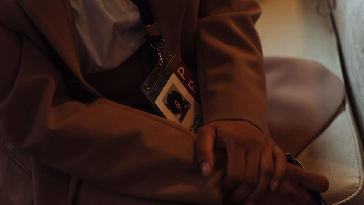 Brown coat-clad person holding an ID badge and pen in a dimly lit setting.