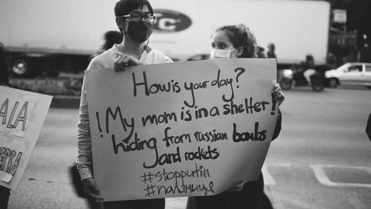 Two masked individuals hold a protest sign about shelter concerns amidst a street scene.