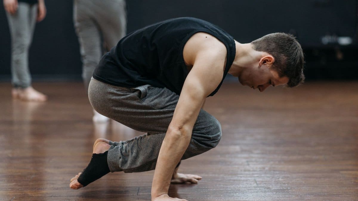 A man in a black tank top practices balance on a wooden floor in a dance studio.