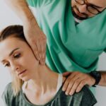 Confident doctor wearing uniform and eyeglasses gently doing therapeutic massage on calm female patients neck and stretching stiff neck muscles