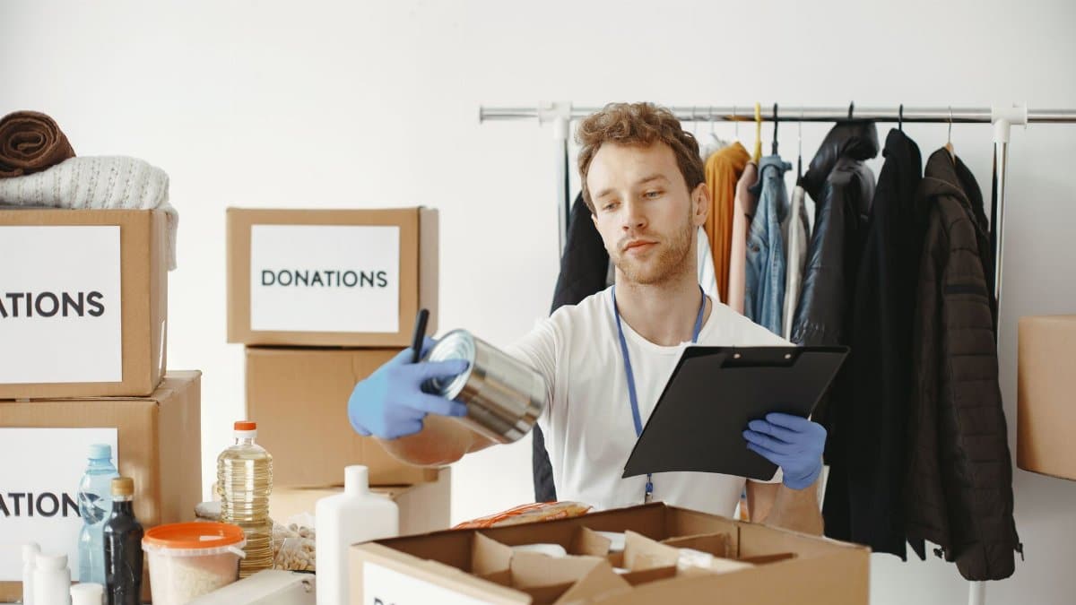 A volunteer organizes food and clothing donations in a community center, maintaining records on a clipboard.