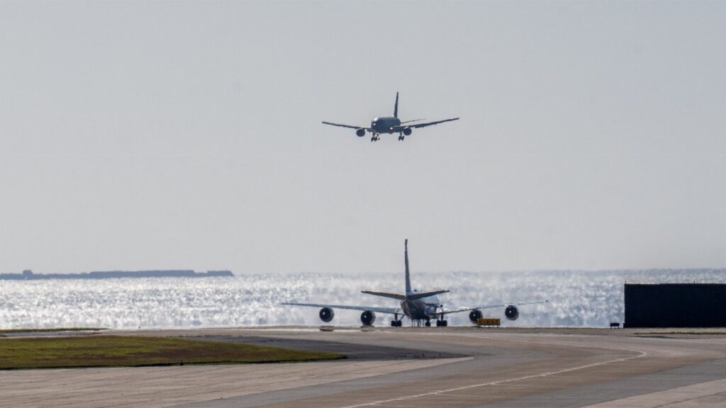 Two airplanes on a sunny runway by the sea, one landing and the other on ground.