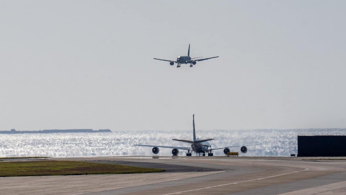 Two airplanes on a sunny runway by the sea, one landing and the other on ground.