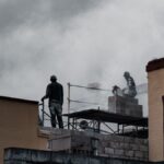 Workers on a multi-storey building site using tools amidst a cloudy backdrop.