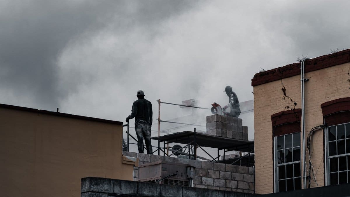 Workers on a multi-storey building site using tools amidst a cloudy backdrop.