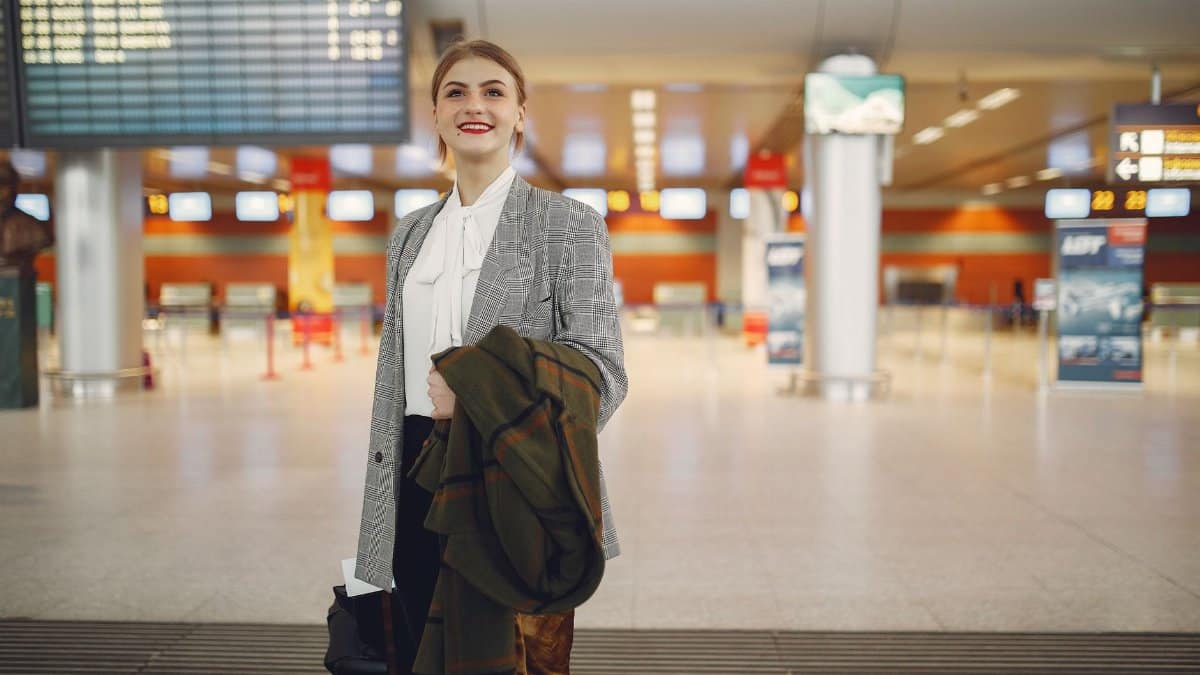 Smiling female manager in elegant wear checking schedule of flights in airport while standing with suitcase and coat looking up