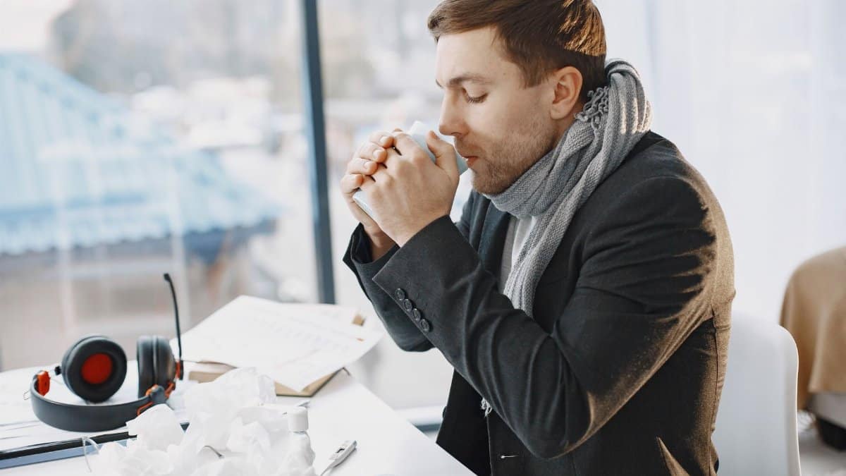 A man in a black suit and scarf drinks coffee in a bright office, suggesting he's unwell.