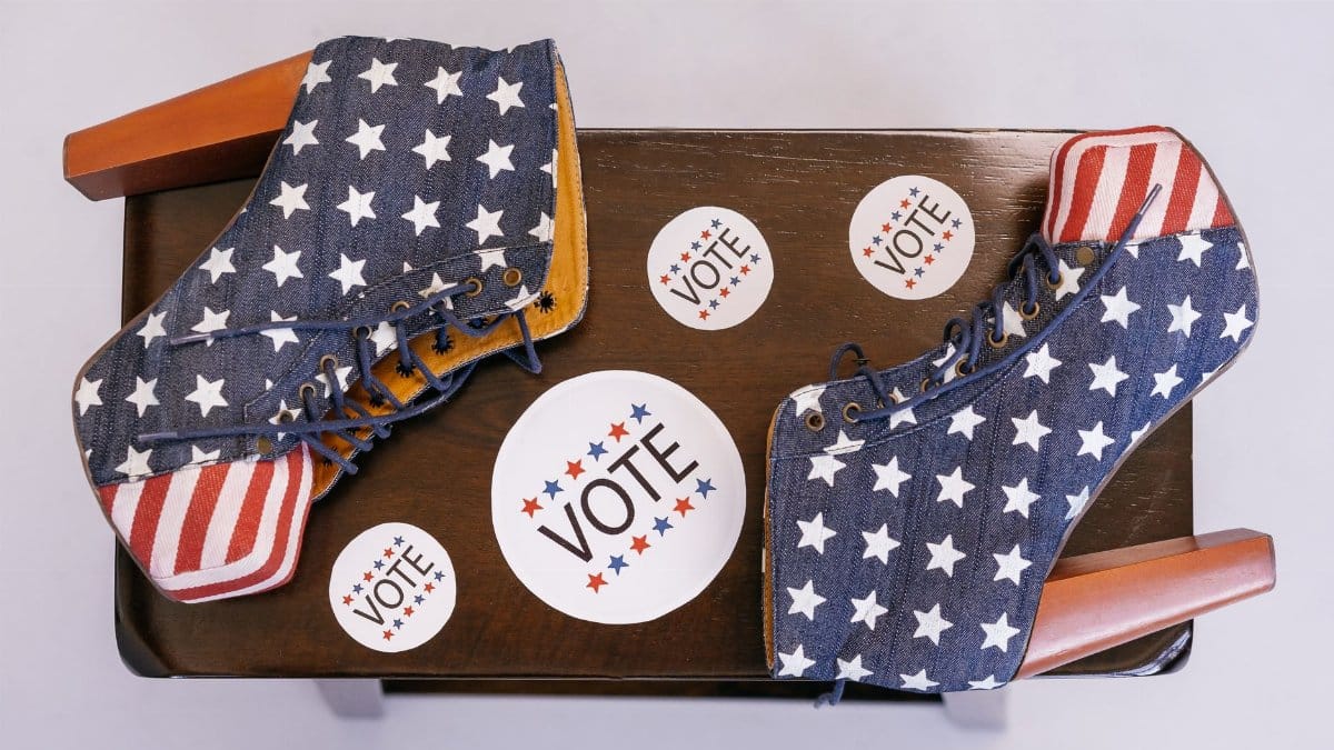 High heel shoes with stars and stripes, surrounded by voting campaign stickers on a wooden surface.