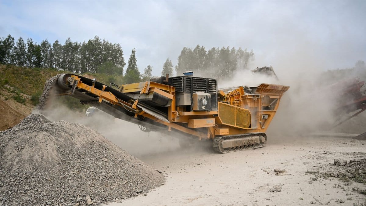 Heavy-duty crusher machine processes materials in dusty quarry amidst greenery.