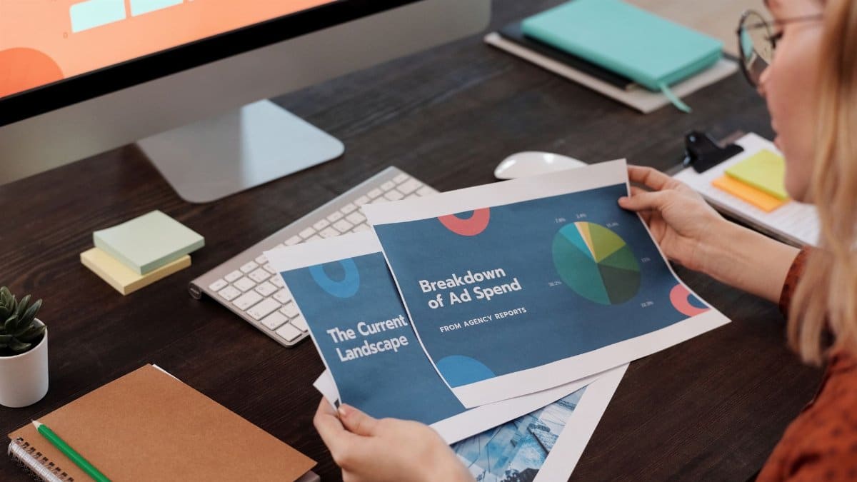 A businesswoman reviews marketing documents with charts at her desk, featuring a computer and office supplies.