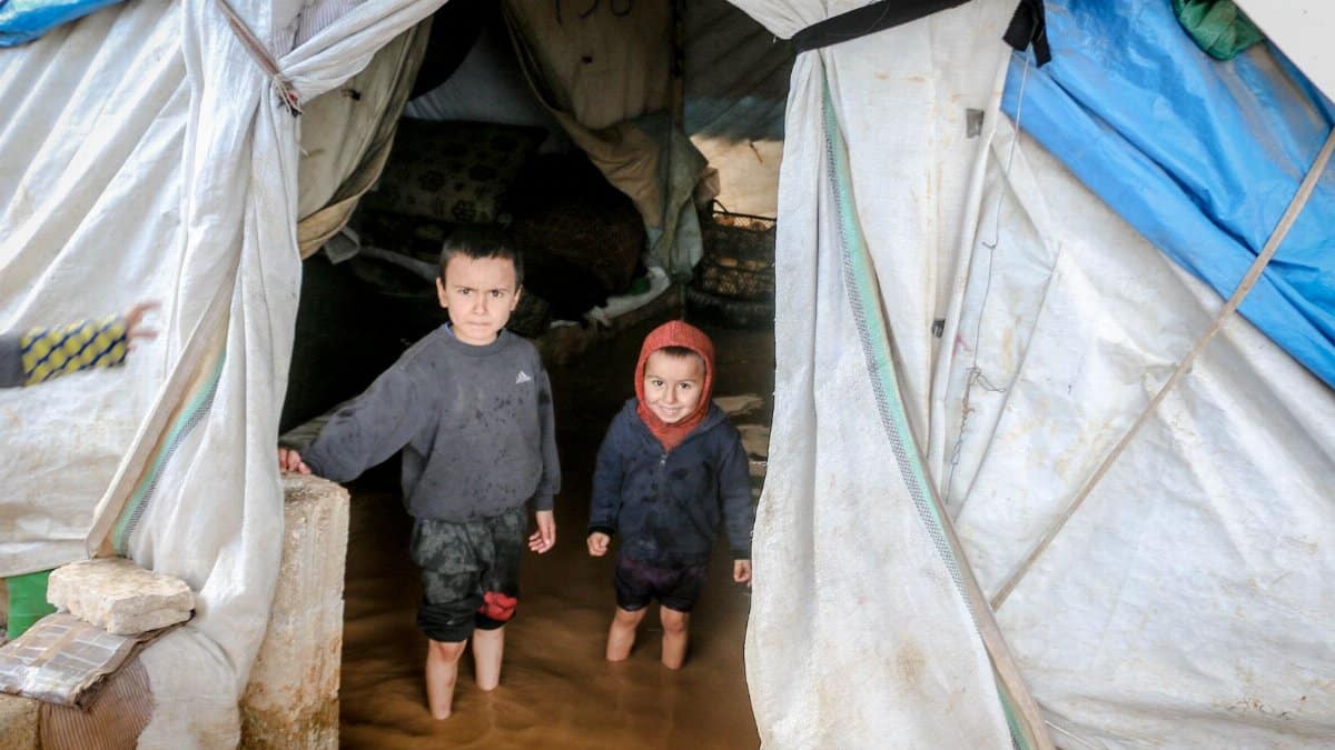Two young children stand in a flooded tent in Idlib, Syria, highlighting the hardships in refugee camps.