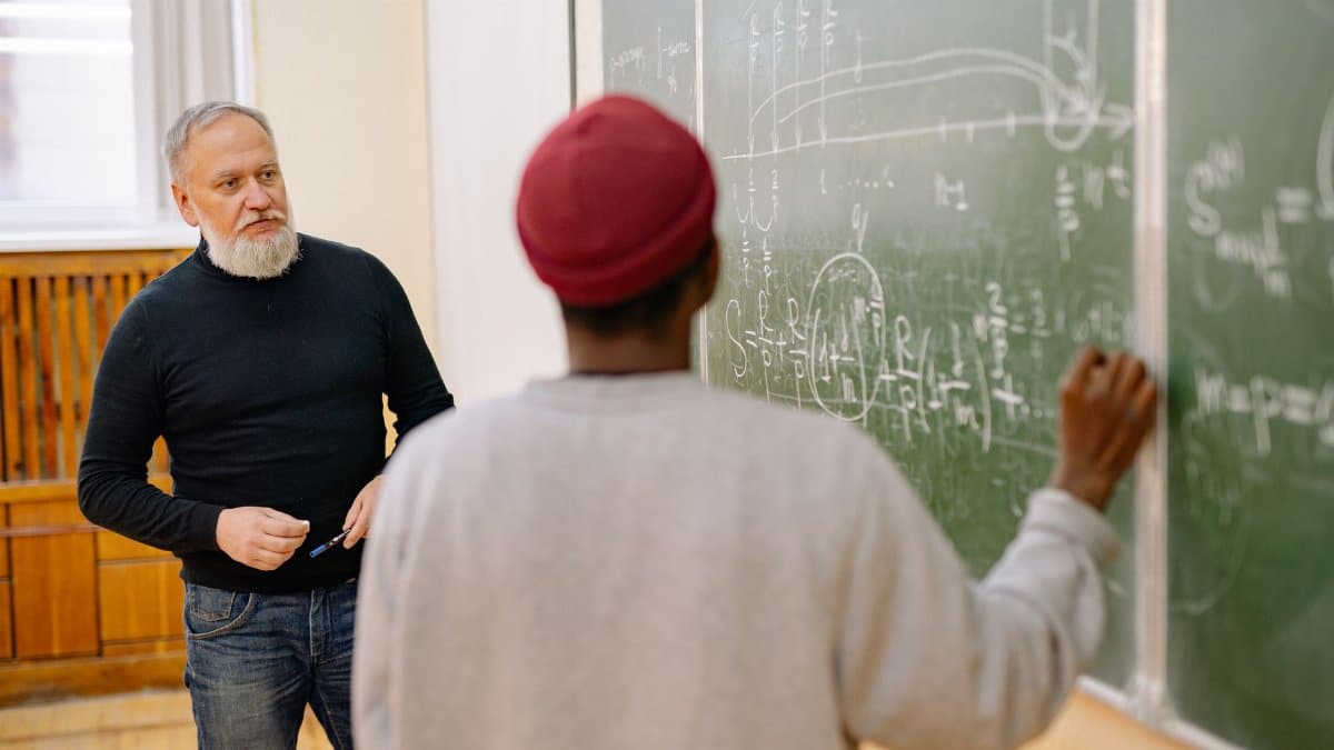 Educator assisting student during a math lesson at a blackboard in a classroom setting.
