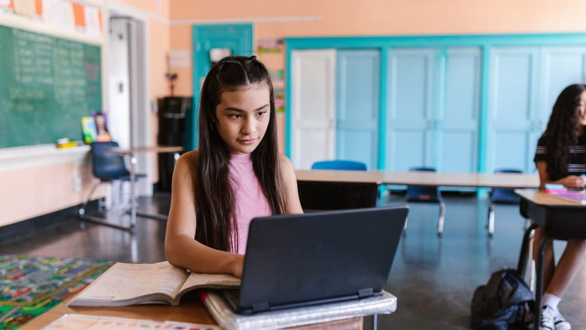 A focused young girl using a laptop for schoolwork in a bright classroom environment.