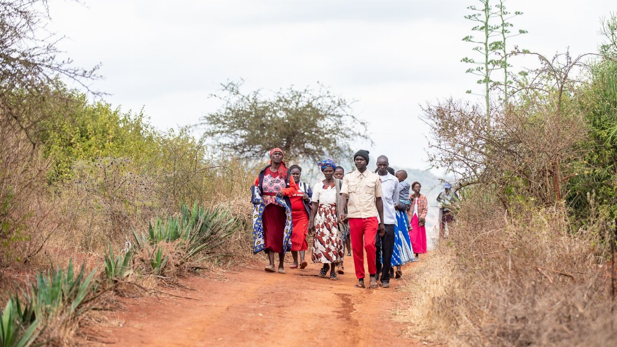 A diverse group of people walking along a rural dirt road surrounded by trees and bushes.