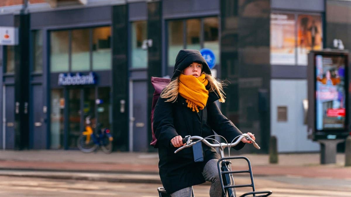 A woman rides a bicycle on an Amsterdam street, wearing a hoodie and scarf. Urban winter scene.