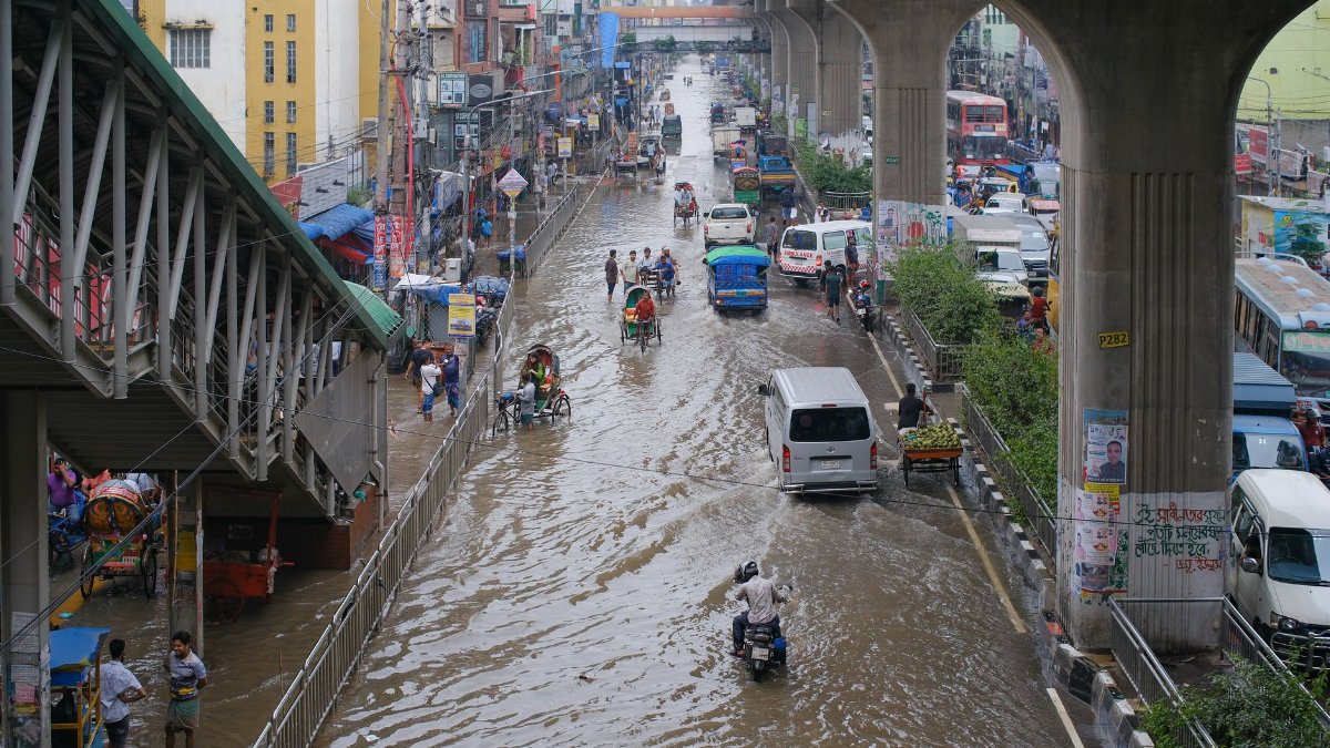 Urban street flooded with vehicles and pedestrians navigating water under elevated metro tracks.