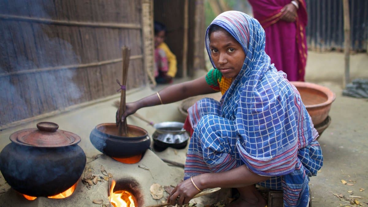 A woman in a blue sari uses a clay stove outdoors in rural Bangladesh, symbolizing traditional cooking methods.