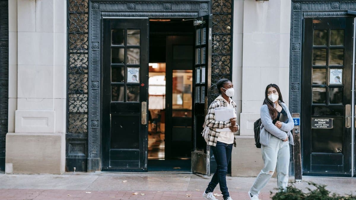 Positive young multiracial female friends in casual wear and protective masks carrying folders and chatting while strolling together on street