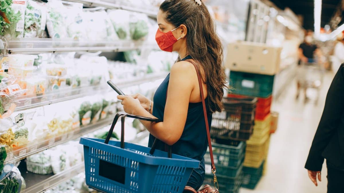 A woman wearing a mask shops for fresh produce while using a smartphone in a grocery store.