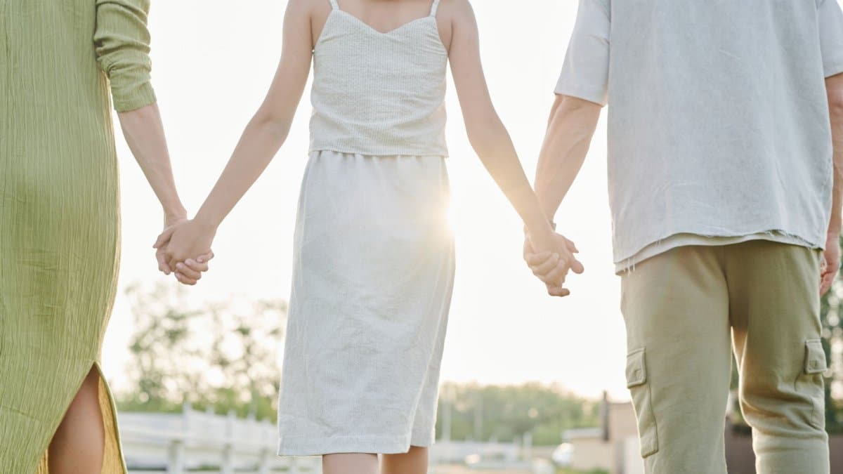 Three people holding hands, walking outdoors in bright sunlight, symbolizing togetherness.