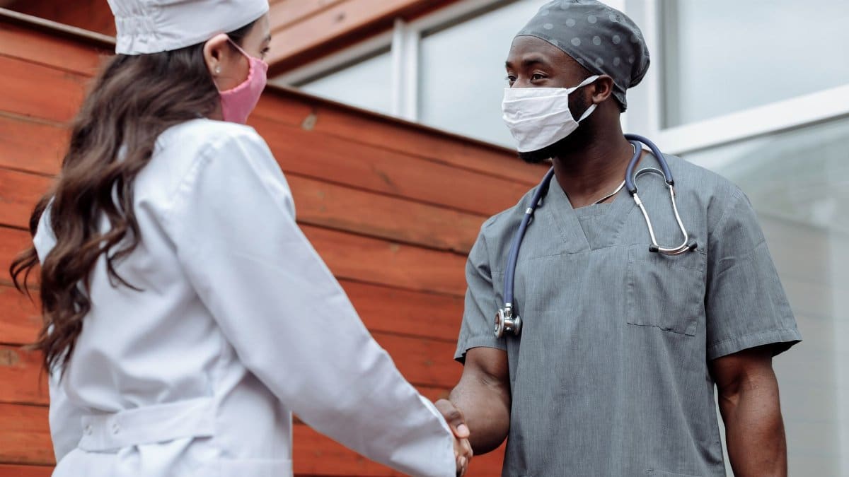 Two healthcare workers in face masks shaking hands outdoors, promoting teamwork and collaboration.