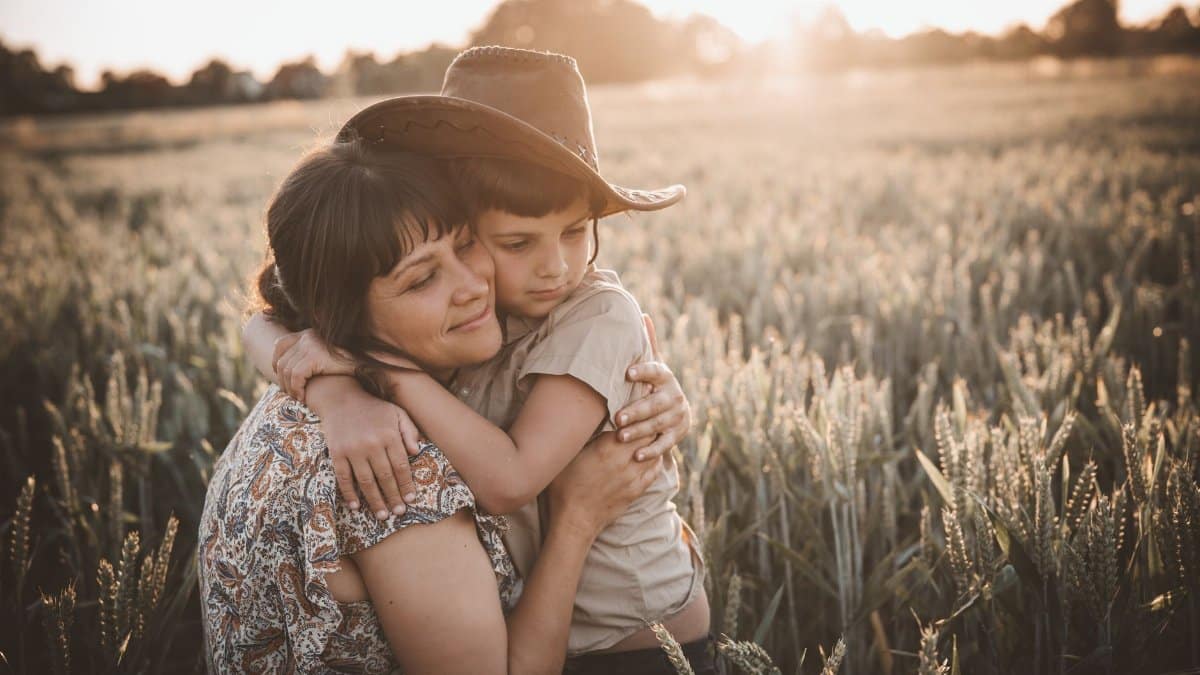 Gentle mom with closed eyes hugging son wearing cowboy hat in rural wheat field at sunset