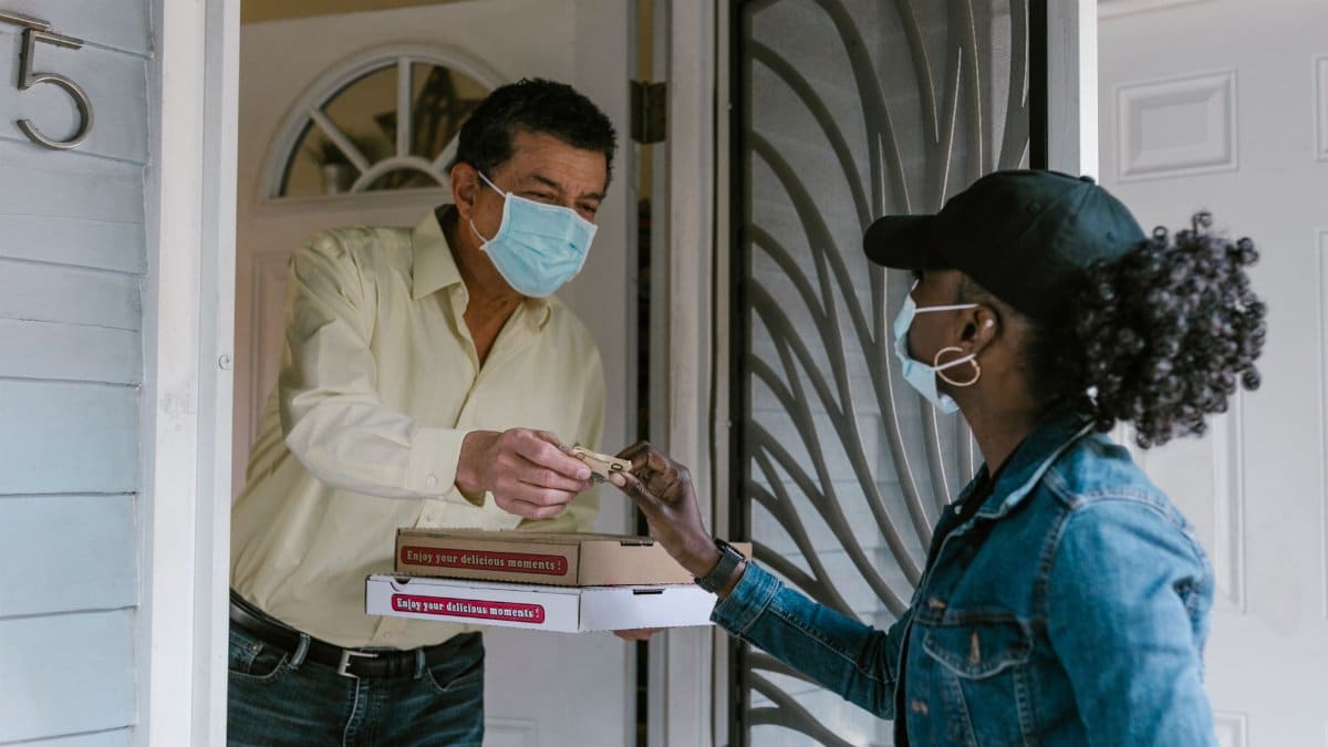 A delivery person hands a pizza to a masked customer at the door, showcasing safe delivery services.