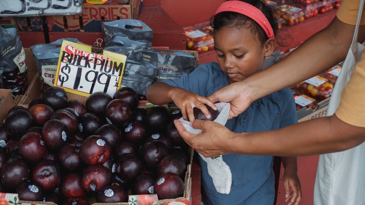 Positive Asian girl putting ripe plums into bag while buying fruits with mother in local street bazaar