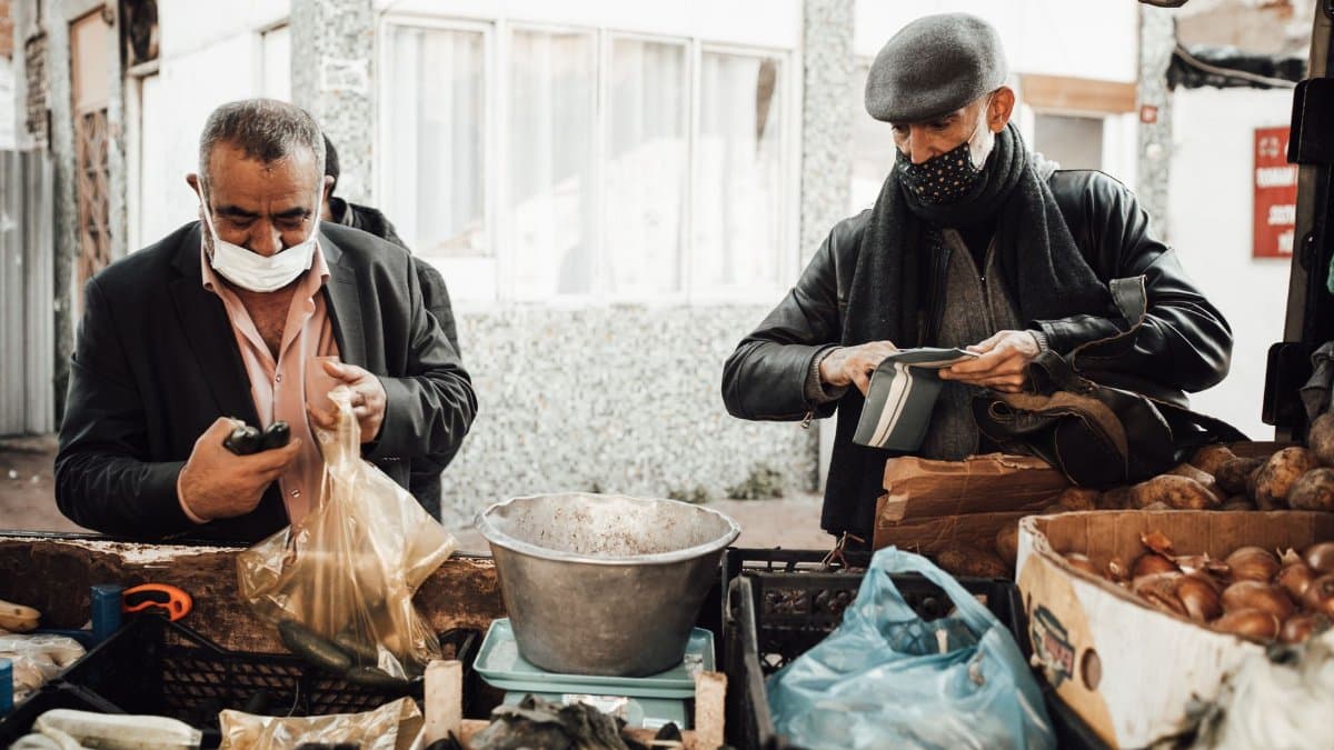 Multiracial male in outerwear and protective masks standing near stall with different products in boxes selling food to buyer in local street market